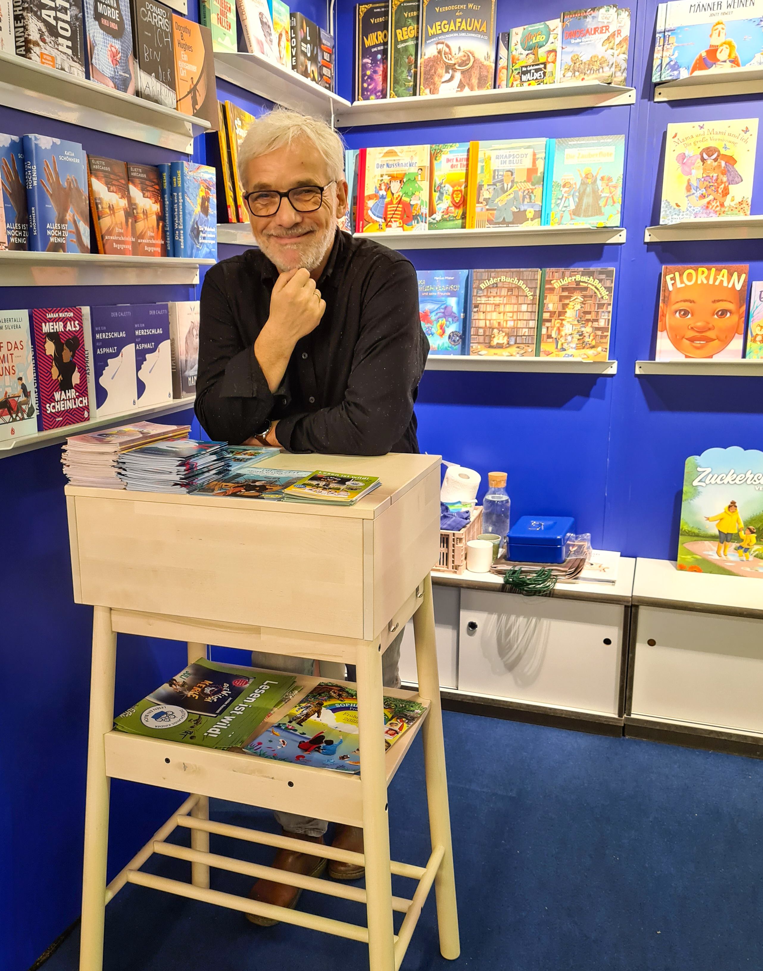 Ein lächelnder Mann lehnt an einem Pult vor einem Bücherregal und stützt seinen Kopf auf der rechten Hand ab.