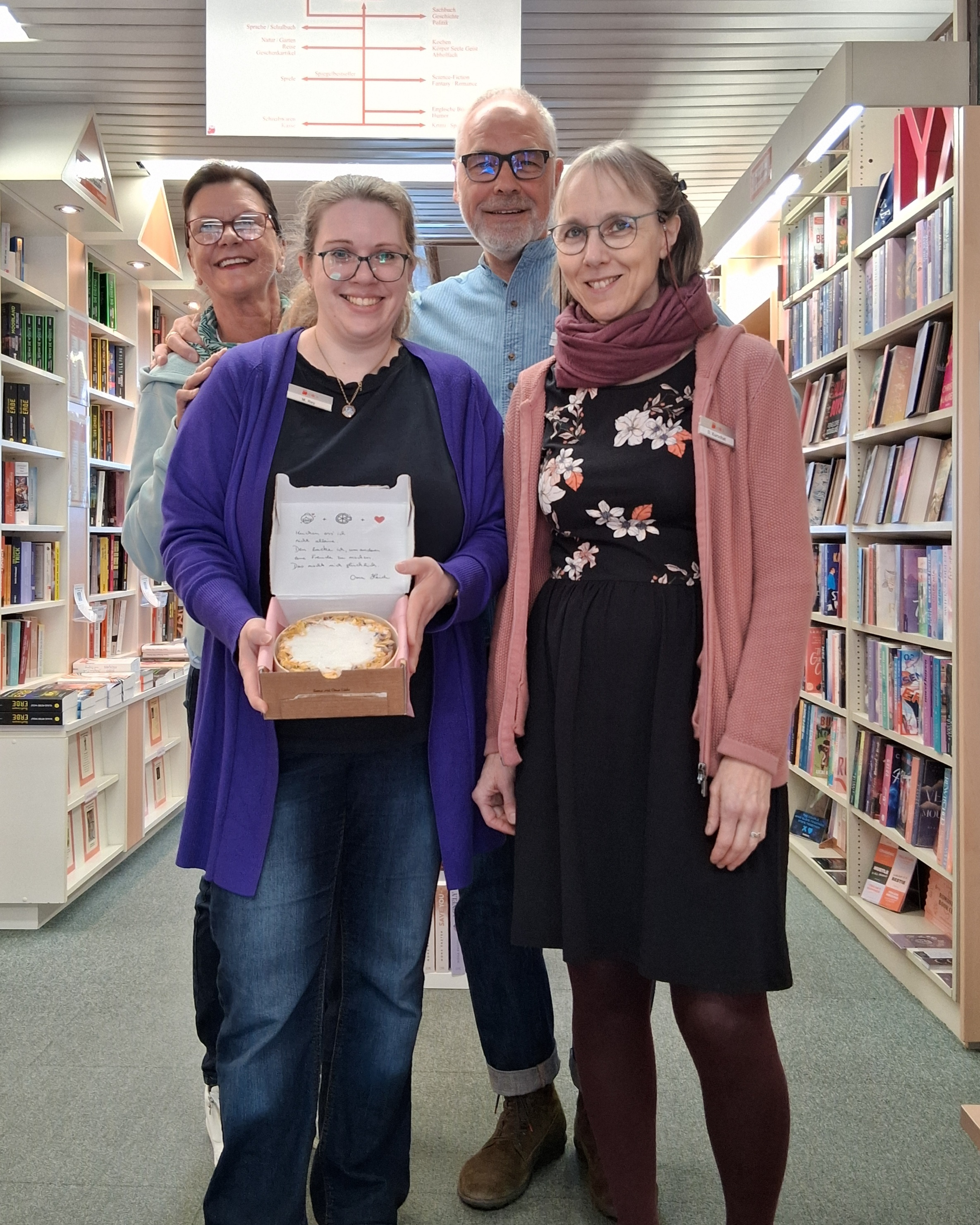 Vier personen in einem Gang einer Buchhandlung, wobei eine frau vorne einen Kuchen in der Hand hält.