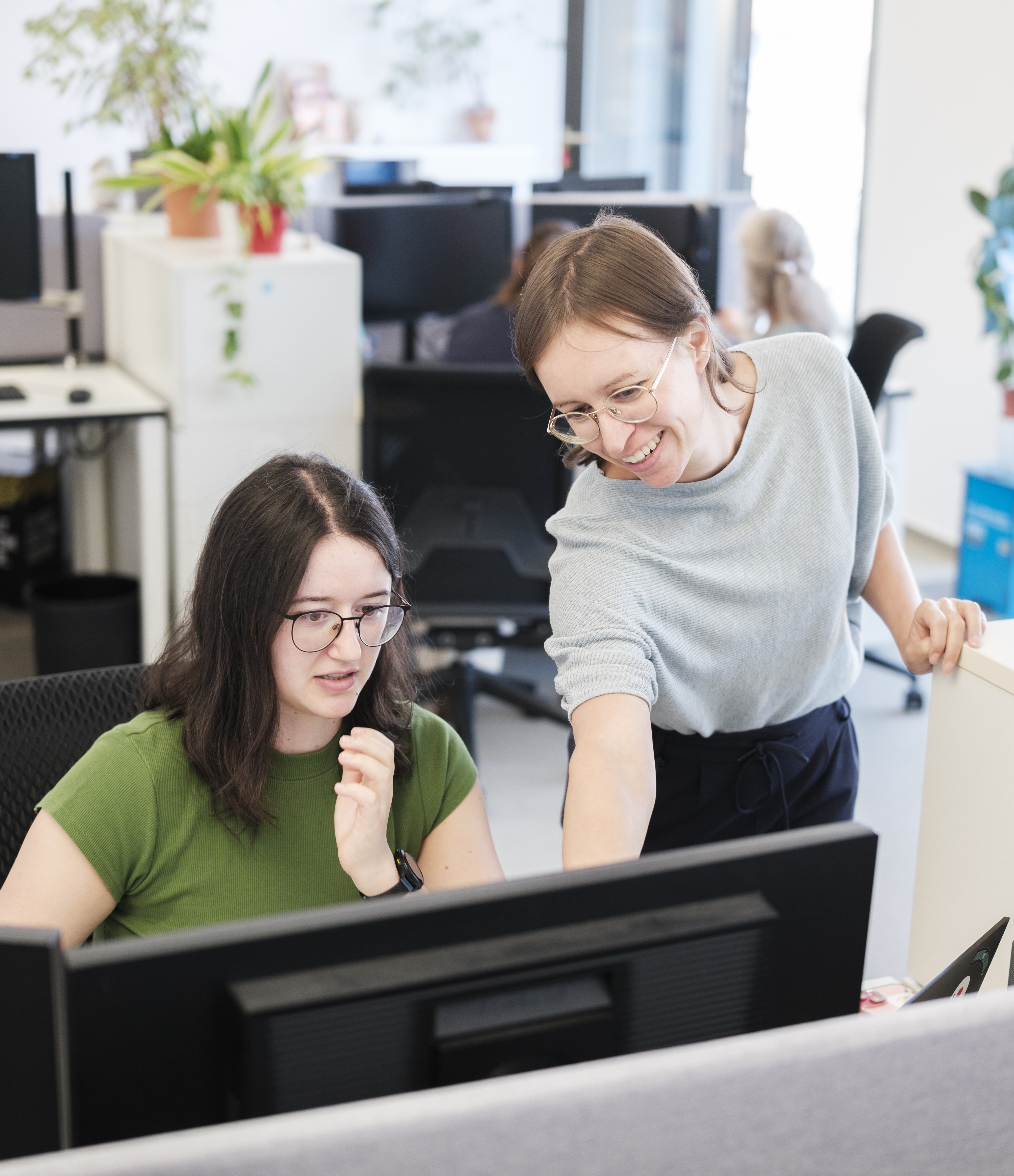 Eine Frau schaut auf einen PC-Bildschirm eine andere steht rechts neben ihr und zeigt auf etwas auf dem Bildschirm.