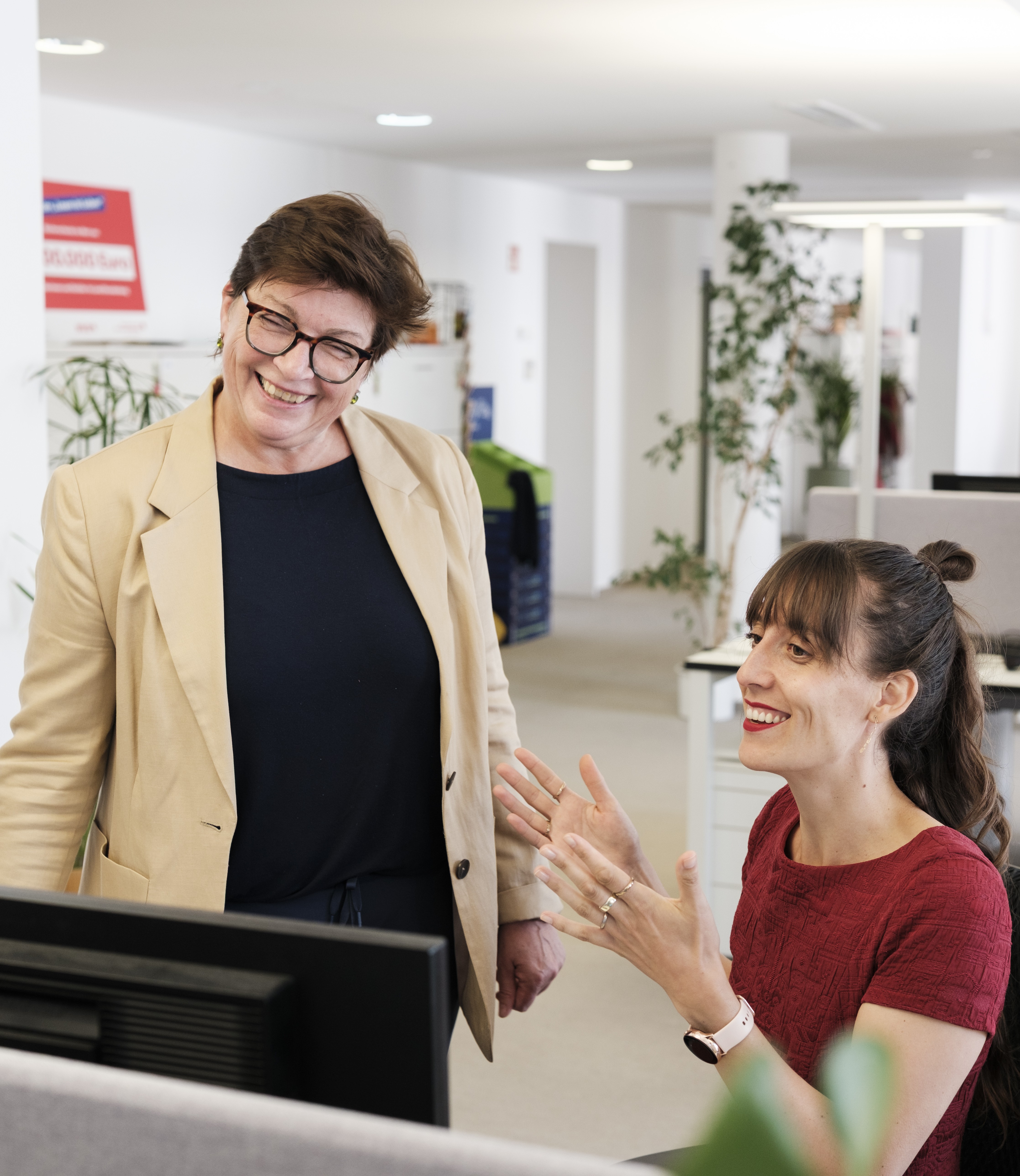 Zwei Frauen sind vor einem Computer am Schreibtisch und lachen.