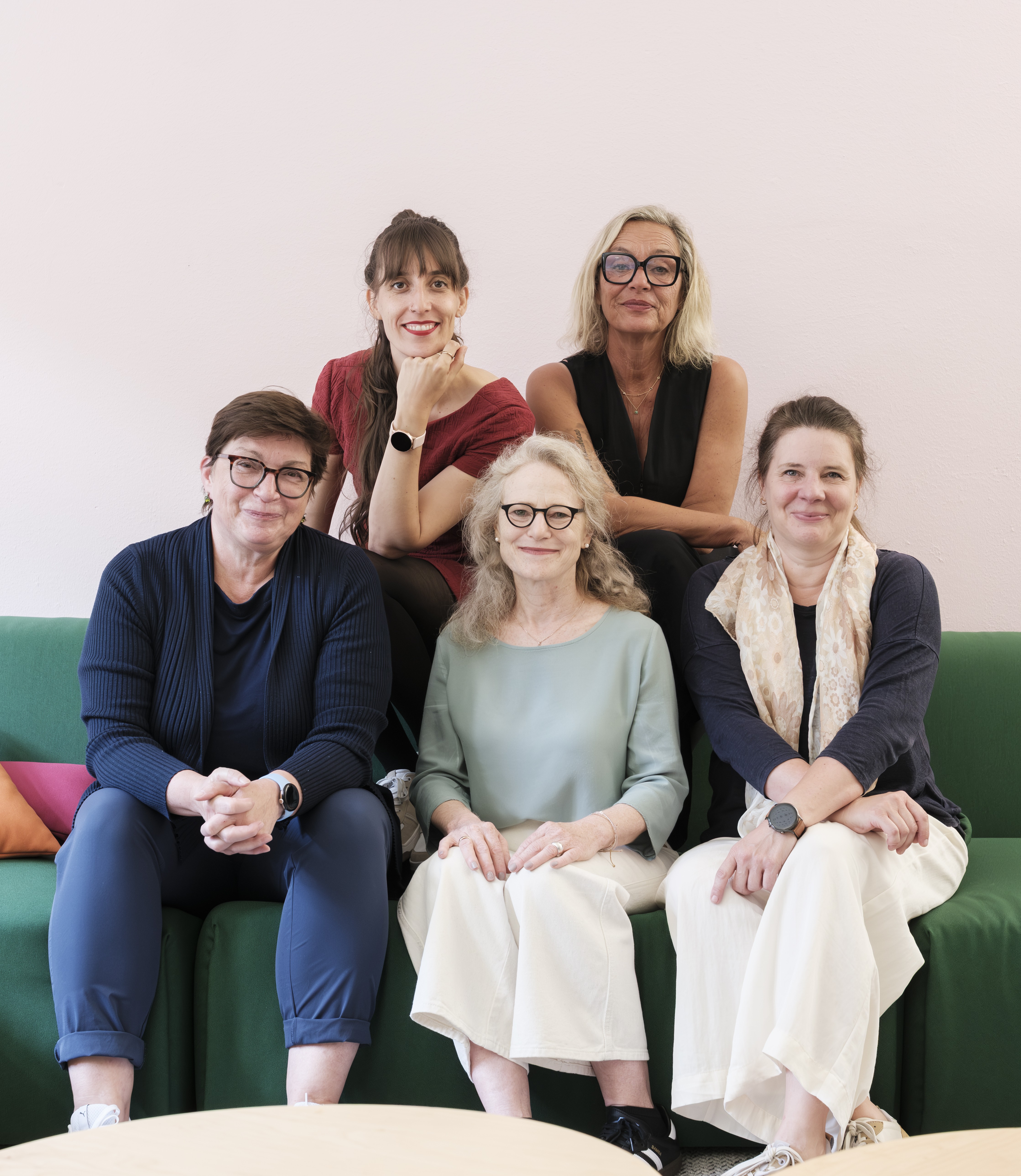 Fünf Frauen sitzen auf einer Couch und lachen in die Kamera.