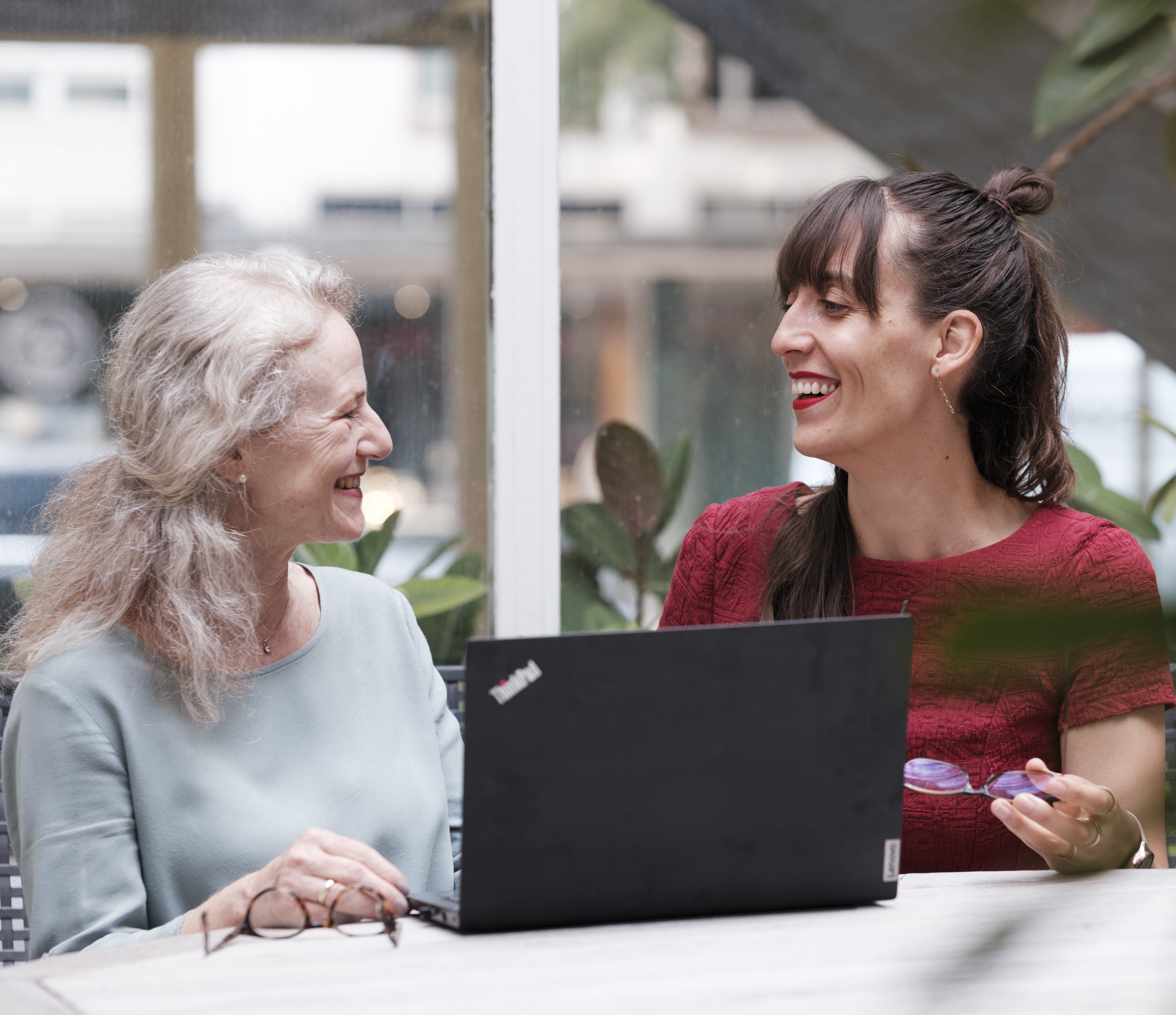 Zwei lachende frauen schauen sich an und sitzen gemeinsam vor einem offenen Laptop am Tisch.